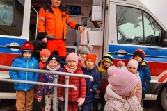 Dzieci stoja pod karetką pogotowia, w tle widać ratownika medycznego. 