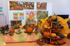 Przedszkolna sala zabaw. Jesienny Kącik przyrody . Poukładane na niskiej szafce kosze z jesiennymi darami ; dynie; orzechy, jabłka , kolorowe kwiaty. Po prawej stronie półka w kształcie słonka z jesiennymi darami i wiklinowe kosze z kasztanami i ozdobnymi dyniami . W tle tablica z licznymi ilustracjami o tematyce jesiennej .