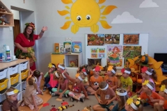 Sala przedszkolna dzieci ubrane na kolorowo z opaskami z kolorowym listkiem na głowach siedza przed tablicą Na podłodze rozrzucone kolorowe liście kasztany i żołędzie. Dzieci zwrócone w stronę drzwi machają w kierunku Pani Jesieni.