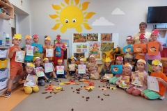 Sala przedszkolna . Dzieci ubrane na kolorowo ustawione przed tablicą trzymaja przed sobą kolorowe rysunki głównie – kolorowanki konturowe grzybów; muchomora i borowika , oraz wycinanki ; kosz z grzybami .