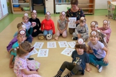 Grupa dzieci wraz z wychowawczynią siedzą na podłodze w kole, na środku leży kilka kartek z jeżami, dzieci mają na głowie opaski z jeżem.