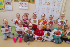 Dzieci siedzą na podłodze w dwóch rzędach, każde z nich trzyma w ręku wyklejonego z plasteliny jeża. Za dziećmi tablica z ciekawostkami o jeżach.