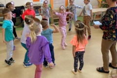 Grupa dzieci stoi rozproszona na sali, dziewczynka w środku pokazuje figurę, inne dzieci robią to samo.