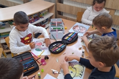 Przy stoliku przedszkolnym siedzi pięcioro chłopców w kolorowych strojach. Przed nimi leżą prace plastyczne z sylwetami jeży, piórniki i kleje. W tle widać kolejny stolik i siedzących przy nim chłopców. Jeden z nich ubrany na niebiesko stoi obok stolika.