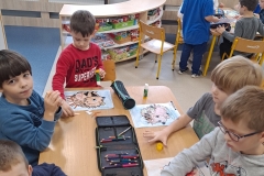 Przy stoliku przedszkolnym siedzi pięcioro przedszkolaków. Dziewczynka i chłopiec mają na sobie białe bluzy z nadrukiem, pozostali chłopcy ciemne bluzy. Dzieci kolorują sylwety jeży na kartkach, wyklejają im kolce wycięte z papieru kolorowego. Na stoliku leżą otwarte piórniki z kredkami i kleje. Z lewej strony widać szafkę             z układankami, w tyle drewniane osłony na kaloryfer.