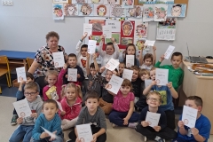 W sali przedszkolnej na podłodze przed ścianą siedzą dzieci w kolorowych strojach. Trzymają w dłoniach białe kartki  - dyplomy z sylwetą jeża i nieczytelnym zapisem.    W tle tablice korkowe z planszami liter i cyfr oraz pracami plastycznymi                         z wizerunkiem jeży. Za dziećmi z lewej strony siedzi nauczycielka.