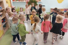 Dzieci poruszają się po kole, trzymając się za ręce. Jedna dziewczynka w żółtej bluzce stoi w środku koła. W rękach trzyma maskotkę jeżyka.