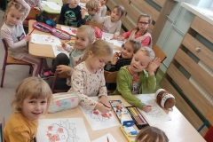 Grupa dzieci siedzi przy 3 stolikach. Przed nimi leżą kartki papieru i kredki. Dzieci kolorują jeża, który na kolcach ma jabłka i gruszki, a pod nogami liście.