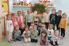 9 dzieci stoi w półkolu. Pomiędzy nimi na środku ustawiona sztaluga, a na niej jeż wykonany z zielonych gałązek i kolorowych liści. Pozostałe 10 dzieci siedzi przed nimi na podłodze.