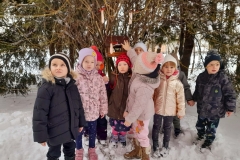 Grupa dzieci stoi przed karmnikiem, na gałązkach widać zawieszoną słoninkę.