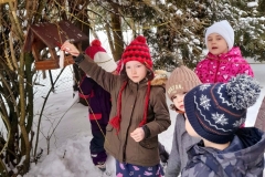 Dziewczynka zawiesza na gałązce drzewa słoninkę, wokół karmnika stoją inne dzieci.