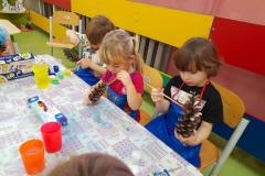 Chłopiec i dziewczynka siedzą przy stole, malują farbami szyszki.