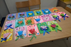 Na stoliku leżą kolorowe kartki w kształcie prostokąta. Do naklejonego koszyczka włożone są kwiatuszki w różnych kolorach. Każda laurka udekorowana jest wstążeczką.