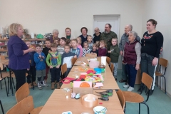 Grupa dzieci i dorosłych stoi na tle białej ściany. Pani w fioletowym swetrze trzyma w rękach koszyk z cukierkami. Na stołach leżą kolorowe kartki, klej, nożyczki.