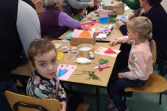 Dzieci i 2 panie siedzą przy stole. 5 dorosłych pochyla się nad nimi. Jedna dziewczynka odwraca głowę do tyłu. Na stoliku leżą kolorowe kartki, klej, nożyczki.