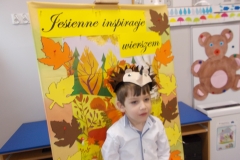1. Na tle jesiennej makiety z kolorowych kartonowych liści stoi widoczny do połowy chłopiec w białej koszuli. Na głowie ma tekturową czapkę - jeża. Na makiecie widoczny jest czarny napis na żółtym tle Jesienne inspiracje wierszem. W tle widoczna tablica z sylwetą misia oraz plansze z cyframi.
