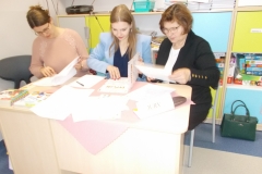 Trzy kobiety siedzą za stolikiem i przeglądają dokumenty. Z przodu po lewej stronie widoczny jest napis: Jury drukowanymi literami, obok leżą dyplomy i książki oraz różne karty. Kobieta siedząca z prawej strony ubrana jest w biała bluzkę, ciemny żakiet i spódnicę. Nosi okulary. Ta     w środku ma granatową bluzkę i jasny niebieski żakiet. Kobieta po lewej stronie ma lekko różową bluzkę i również nosi okulary. W tle widoczna ciemna torebka i szafki przedszkolne.