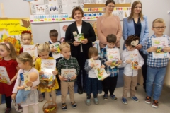 3. W sali przedszkolnej stoją trzy kobiety  a przed nimi  grupa dzieci ubranych w kolorowe stroje. Dzieci trzymają w ręku karty z widocznym napisem: ,, Dyplom’’, reszta treści jest nieczytelna. Mają też małe książki z kolorowymi okładkami. Tytuły są nieczytelne. Przed jedną z dziewczynek stoi na podłodze wiklinowy koszyk z owocami.