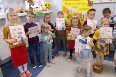4. Grupa dzieci ubrana w kolorowe stroje stoi w sali przedszkolnej. W ręku trzymają dyplomy o treści: Dyplom dla ( imię i nazwisko dziecka) za zajęcie I miejsca w konkursie recytatorskim Jesienne inspiracje wierszem. Podpisy członków Jury. Data: 29. 11. 2022 r. Trzymają też dodatkowo książeczki. Dobrze widoczny tytuł jednej z nich to Samochwała. Dzieci stoją na tle jesiennej makiety z kolorowymi kartonowymi liśćmi oraz czarnym napisem na żółtym tle: Jesienne inspiracje wierszem. Z boku po lewej stronie widoczne okno, na parapecie rozłożona jesienna dekoracja.