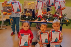 Na stole leżą stroiki świąteczne  za nimi stoi 5 dzieci z dyplomami i nagrodami, na podłodze siedzi troje dzieci z dyplomami I nagrodami.