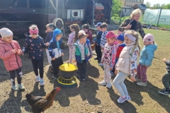 Dzieci stoją w półkolu na ziemi. Wśród nich pani w ciemnej bluzce. Przed nimi stoi mały paśnik dla kur. Obok niego jedna brązowa kura.