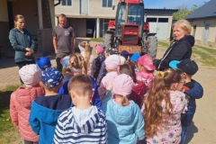 Grupa dzieci w kolorowych strojach stoi na podwórku. Przed nimi pan w brązowym podkoszulku i pani w niebieskiej bluzie, a obok pani w czarnym dresie. Na środku podwórka duży, czerwony traktor.