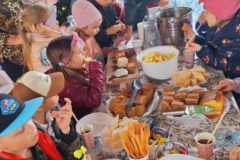 Dzieci siedzą w altance przy drewnianym stole. Z apetytem jedzą przygotowane smakołyki: ciasto, chrupki, cukierki, sery. Piją wodę, soki, herbatę. Pani w niebieskiej bluzie podaje dzieciom talerzyki.