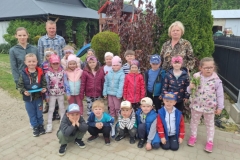 Grupa dzieci w kolorowych strojach stoi na szarej kostce. Przed nimi przykucnęło 5 chłopców. Za dziećmi stoją 2 panie i1 pan w kraciastej koszuli. W tle zielone i czerwone krzewy.