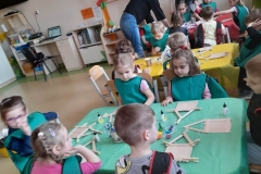 Dzieci siedzą przy 3 stolikach, ķleją domek z patyczków, dwie panie pomagają przy ostatnim stoliku.