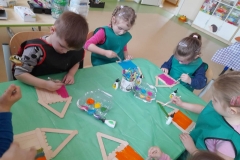 Przy stoliku siedzi 5 dziewczynek, malują farbami domki z patyczków.