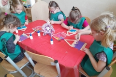 Dzieci siedzą przy 3 stolikach, ķleją domek z patyczków, dwie panie pomagają przy ostatnim stoliku.