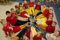 Dzieci w czerwono - żółtych ubraniach siedzą na podłodze w kole. Proste nogi wyciągnięte do przodu łączą się w środku koła. Ręce skierowane do tyłu opierają o podłogę.