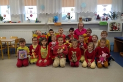 Grupa dzieci z wychowawczynią siedzi na jasnej podłodze. Niektórzy trzymają w rękach maskotki: Kubusia Puchatka, Króliczka, Tygryska, Prosiaczka, Kangurzycy, Sowy. Jeden chłopczyk ma książkę. W strojach całej grupy dominuje kolor czerwony i żółty.