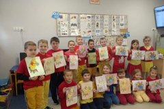 Dzieci w I rzędzie siedzą na podłodze, a w II stoją za ich plecami. Niektórzy trzymają obrazki z Kubusiem Puchatkiem, inni ilustracje przedstawiające mieszkańców Stumilowego Lasu. Za nimi na ścianie wisi godło oraz plansze z literki i cyframi.