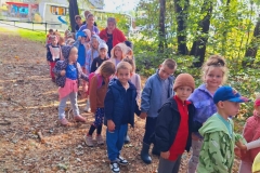 Dzieci ustawione w pary idą leśną drogą.  Za nimi widać zielone liście krzewów i drzew.