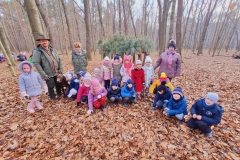 Grupa dzieci ubrana w kolorowe stroje kuca przed paśnikiem. Obok stoi mężczyzna ubrany w zielony strój.  Trzyma przed sobą dużego brązowego psa. Z tyłu stoi pani w zielonym stroju oraz paśnik.