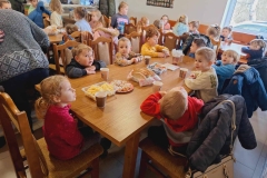 Dzieci siedzą na krzesłach przy stole. Na stole stoją  talerze z rogalikami,  chleb w koszyczku,  kubeczki z herbatą.