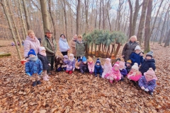 Grupa dzieci ubrana w kolorowe stroje kuca przed paśnikiem. Obok stoi mężczyzna ubrany w zielony strój.  Trzyma przed sobą dużego brązowego psa. Z tyłu stoi pani w zielonym stroju oraz paśnik.