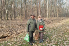 Kobieta i mężczyzna stoją  obok siebie.  Obydwoje ubrani w ciemnozielone stroje. Mężczyzna trzyma w rękach zieloną i brązową torbę,  kobieta czerwony okrągły pojemnik. W tle  drzewa.