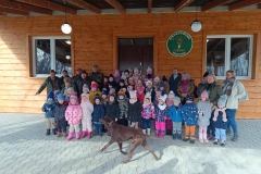 Duża grupa dzieci ubranych w kolorowe  kurtki i czapki stoi w kilku szeregach. Za nimi stoją  osoby dorosłe. Na pierwszym planie  widać  dużego brązowego psa. W tle brązowa ściana  domu z dwoma oknami i drzwiami.