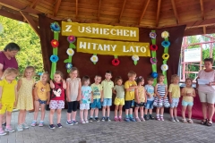 Grupa dzieci ubranych w kolorowe stroje stoją w szeregu. Obok nich dwie panie. Z tyłu na bordowym tle widnieje napis Z uśmiechem witamy lato
