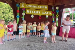 Dwie dziewczynki stoją obok siebie. Przy nich stoi kobieta w jasnej bluzce i krótkich spodenkach. Za nimi stoją dzieci w szeregu.