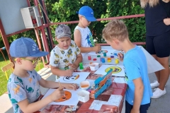 Czterech chłopców stoi przy stoliku. Obok nich pani ubrana w ciemny strój. Dzieci malują farbami.