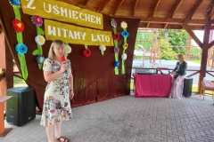 Na zdjęciu widać kobietę ubraną w jasną sukienkę. W tle widoczny napis Z Uśmiechem witamy lato