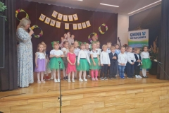 Siedem dziewczynek stoi w rzędzie. Ubrane  w białe  bluzeczki I niebieskie spódniczki. Obok  stoi pięciu chłopców ubranych w jasne koszule  i ciemne spodnie. Za nimi na ciemnym tle kartki i koła.