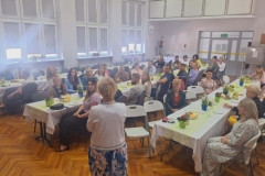W dużej sali stoją  3 rzędy stołów,  a przy  nich panie i panowie ubrani w kolorowe stroje.