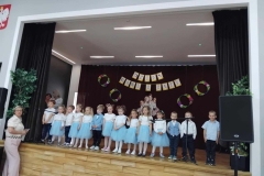 Dzieci ubrane w zielone spódniczki I białe bluzeczki stoją w trzech rzędach. Obok stoi pani w długiej  jasnej sukience. Za nimi na ciemnym  tle widać kartki I kółka.