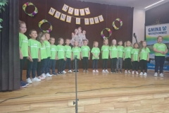 Duża  grupa dzieci stoi, tworząc półkole.  Dziewczynki i chłopcy ubrani są  w zielone koszulki, ciemne getry i spodnie. Za nimi na brązowym tle widać kartki i cztery kółka.