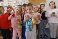 Na tle tablicy z literami stoi grupa dzieci przedszkolnych w kolorowych strojach. Na przodzie stoją chłopiec w jasnym ubraniu z nadrukiem oraz dziewczynka w różowym stroju. W dłoniach trzymają soki marchwiowo - owocowe z napisem na etykiecie:   ,, Smakotek’’. Wszystkie dzieci mają w dłoniach kolorowe kubki na sok.