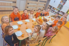 Dzieci siedzą na krzesełkach przy złączanych stolikach . Przed każdym dzieckiem na serwetce biały talerzyk . Obok kubek przeźroczysty z sokiem marchewkowym . Na środku stolika ; talerz z musem marchewkowym, marchewki w koszyku i ciasto marchewkowe.
